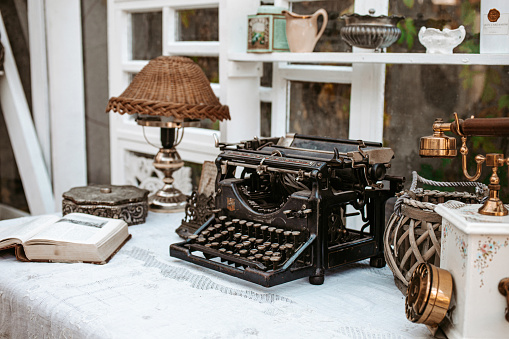 Vintage old fashioned typewriter machine on desk next to window with lamp, books and retro phone in light white shabby chic vintage french Provence country cottage interior room with antique objects