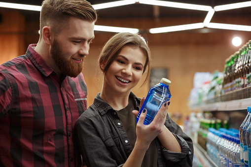 Image of young cheerful loving couple in supermarket choosing water. Looking aside.