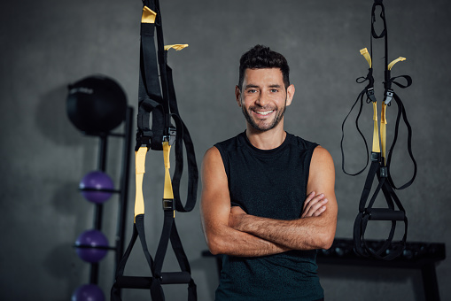 Confident smiling hispanic man looking at camera in sport club. Fitness instructor standing arms crossed in fitness center. Real people in their job. Copy-space