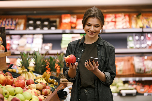 Image of happy young lady standing in supermarket holding apple and chatting by phone. Looking at phone.