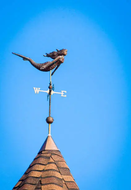 A mermaid weathervane with flowing hair swims atop a domed roofline on Cape Cod against a blue sky.
