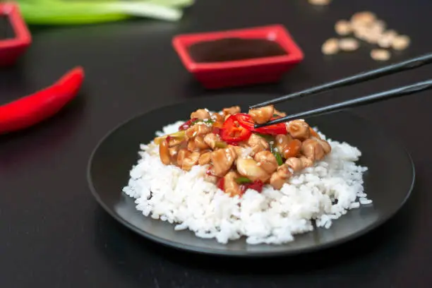 Photo of black Chinese chopsticks hold a piece of kung pao chicken on a plate with rice and a traditional, popular Chinese dish consisting of chicken, peanuts, hot chili pepper, sauces and spices.