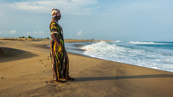 Woman from Ghana in long suit and headdress standing by the beach and looking out over the sea in Keta Ghana West Africa