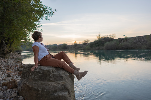 Mature woman relaxes by lakeside at sunset.sitting on rock.