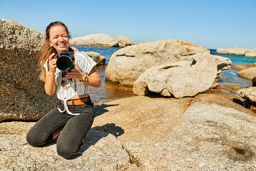 Smiling female photographer sitting at rocky beach and talking pictures with her professional DSLR camera on a summer day