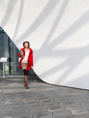 Curly woman in red coat is walking by white wall. Smiling student with laptop. Freelancer with portable device for work. Modern lifestyle.