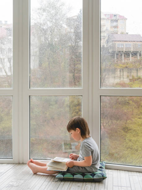 kind sitzt auf dem boden und liest buch. intellektuelle freizeit für kinder, während es draußen regnet. - lesen regen stock-fotos und bilder