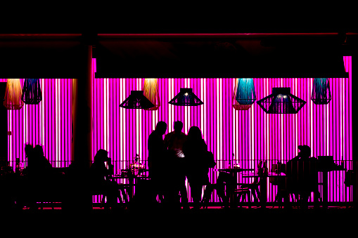 Silhouettes of people sitting in an restaurant, cafe, coffee shop lit with pink light in the city, during the weekend's evening . Photo taken from public street.