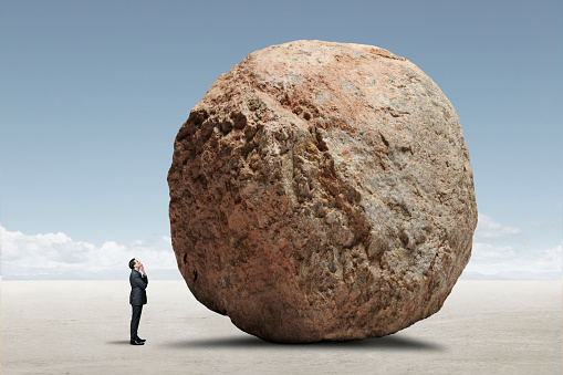 A businessman places his hand under his chin as he looks up at a large boulder that stands as an obstacle to him.