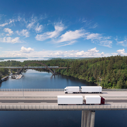 Aerial view of a highway bridge where two trucks with trailers pass by outside Stockholm, Sweden. In the background is railway bridge where a passenger train pass by.