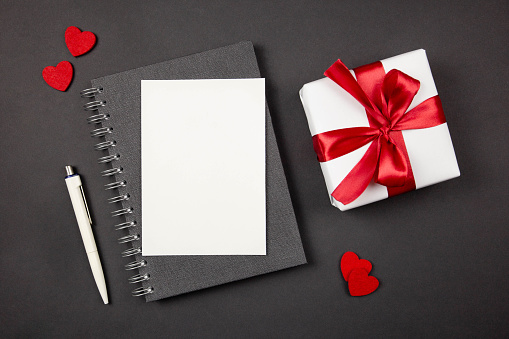 Greeting card mockup and gift box with red ribbon and hearts on dark background. Valentines, mothers, womens day, anniversary and birthday greeting flat lay. Top view. Copy space.