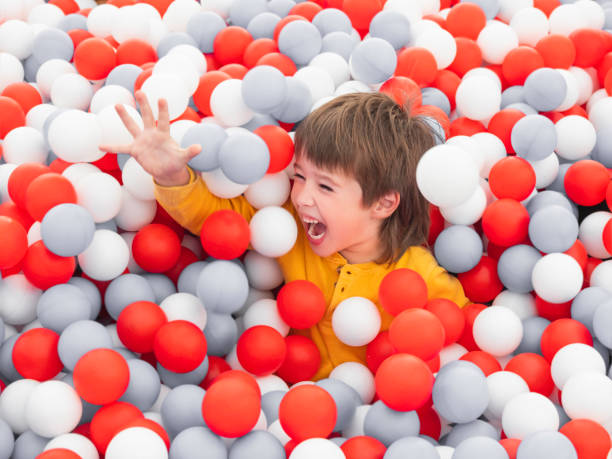 Laughing boy plays in ball pit. White and red plastic balls in dry paddling pool. Leisure activity in playroom at kindergarten. Leisure activity. Active recreation. stock photo