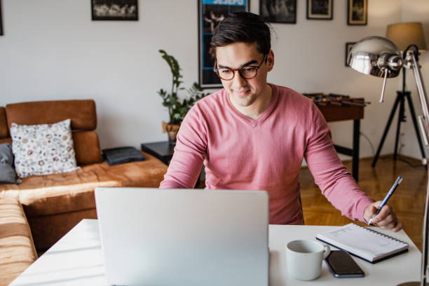 Young man is working on laptop A young man with a laptop at home, he is working and writing notes in a notebook studying stock pictures, royalty-free photos & images