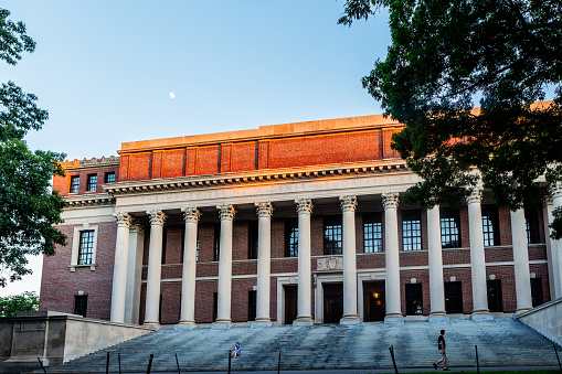 Cambridge, Massachusetts, USA - June 20, 2021: The Harry Elkins Widener Memorial Library (c. 1915) at sunset. The library houses some 3.5 million books in its stacks and is the center­piece of the Harvard College libraries.  It honors 1907 Harvard College graduate and book collector Harry Elkins Widener. Moon and two students visible.