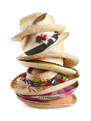 Vertical stack of eight straw hats in a variety of shapes, textures, colors, and sizes, trimmed with ribbons, feathers, and raffia. Isolated on white background, vertical format.