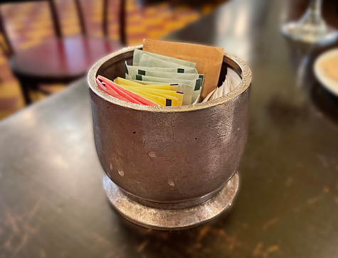 A round, metallic sugar container.   A variety of sweetner envelopes inside on a table.