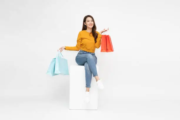 Photo of Young Asian woman holding shopping bags and sitting on white box isolated on white background