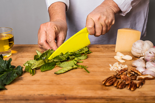 Cropped view of an unrecognizable adult woman cutting basil with a plastic knife on a wooden table for the preparation of a pesto sauce.