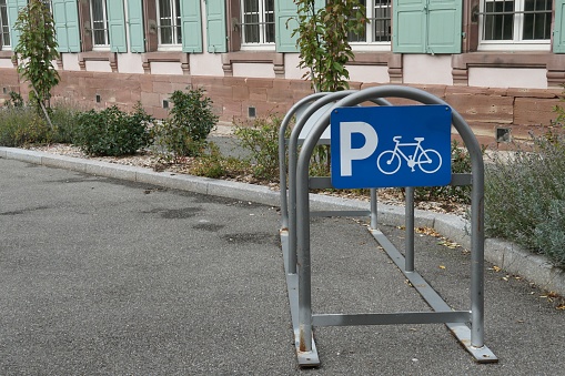 Arch metal frame for parking bicycles with blue sign containing letter P and pictogram of bicykle situated on the street near green house and pavement with vegetation of plants, grass and bushes.