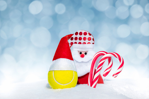 Christmas card with Santa Claus, candy canes, tennis ball in red Santa hat on natural snow on gentle blue background with bokeh. Happy New Year and Christmas concept