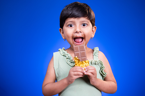 Excited Girl kid with large eyes open, enjoying eating chocolate on blue studio background - concept of unhealthy food consumption and childrens love on chocolate.