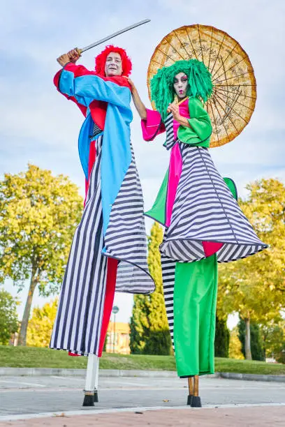 Full length of creative talented performers with face art in colorful costumes and wigs standing on stilts in park with Asian umbrella and sword during show, on sunny day