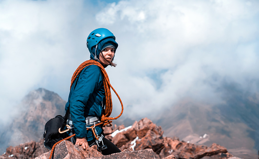 A young cheerful girl in a turquoise helmet, windbreaker and with a red climbing rope sits on the top of the mountain higher than clouds, the traveler successfully climbed the mountains.