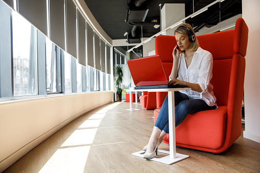 Wide shot of diligent young businesswoman sitting in a red armchair in a modern, open plan office space and video chatting with a colleague via laptop.