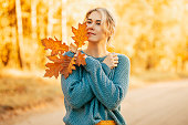 Portrait of young, happy woman looking hopefully into distance, dressed in blue knitted sweater, holding autumn leaves against the background of a road in an autumn park.