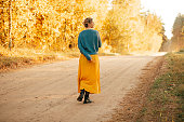 young woman walks along road among autumn forest wearing long yellow skirt and blue sweater, rear view, enjoy solitude, tranquility, silence