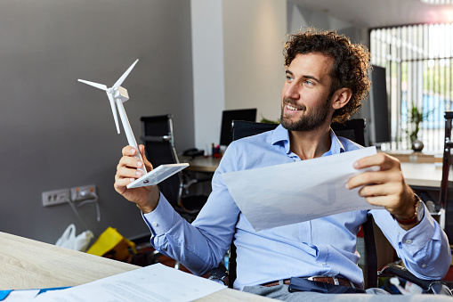 Happy male entrepreneur making plans for sustainable energy while holding wind turbine in the office.