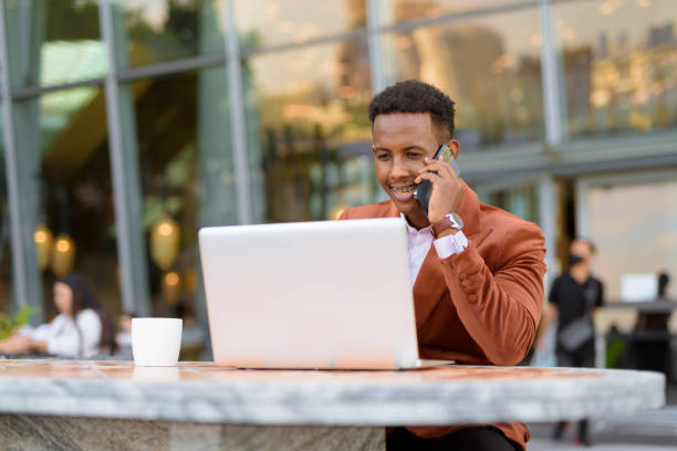 istock Happy African businessman outdoors in coffee shop using laptop computer while talking on mobile phone 1353964279