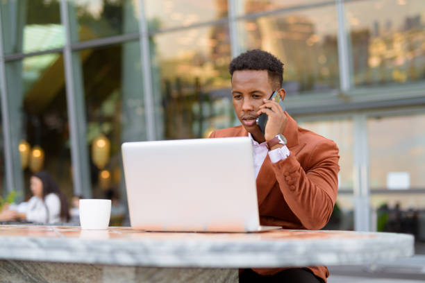 istock African businessman outdoors in coffee shop using laptop computer while talking on mobile phone 1353965106