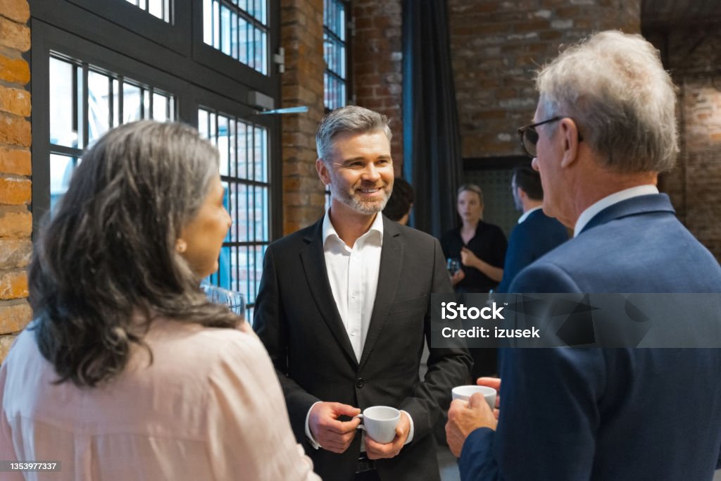 Business people during seminar Group of mature entrepreneurs discussing during business seminar. Smiling businesswoman and businessmen standing in convention center in the evening. Coffee Break Stock Photo
