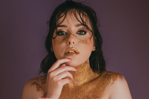 Portrait of a beautiful young woman covered in golden glitter, studio shot in front of purple background