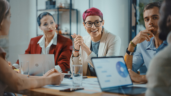 Office Meeting in Conference Room: Beautiful Specialist with Short Pink Hair Talks about Firm Strategy with Diverse Team of Professional Businesspeople. Creative Start-up Team Discusses Big Project