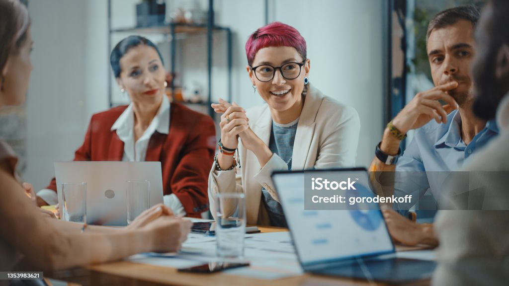 Office Meeting in Conference Room: Beautiful Specialist with Short Pink Hair Talks about Firm Strategy with Diverse Team of Professional Businesspeople. Creative Start-up Team Discusses Big Project Teamwork Stock Photo