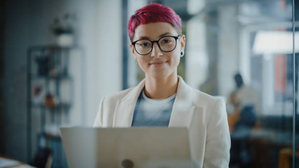 현대 사무실 : 짧은 분홍색 머리 서, 노트북 컴퓨터를 들고, 카메라를보고, 매력적으로 미소 아름다운 정통 전문가의 초상화. 설계, 데이터 분석, 계획 전략 작업 - women computer business laptop 뉴스 사진 이미지