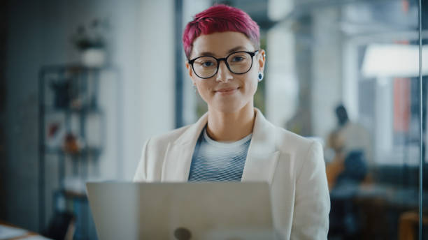 modern office: portrait of beautiful authentic specialist with short pink hair standing, holding laptop computer, looking at camera, smiling charmingly. working on design, data analysis, plan strategy - common imagens e fotografias de stock