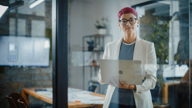 modernes büro: porträt einer schönen authentischen spezialistin mit kurzen rosa haaren, die steht, einen laptop hält, in die kamera schaut und charmant lächelt. arbeiten an design, datenanalyse, planungsstrategie - charmingly stock-fotos und bilder