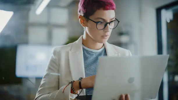 Photo of Modern Office: Portrait of Beautiful Creative Specialist with Short Pink Hair Standing, Holding Laptop Computer. Working on App Design, Data Analysis, Plan Strategy for Social Media Disruption