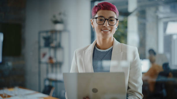 ufficio moderno: ritratto di bella specialista creativa con capelli rosa corti in piedi, tenendo in mano computer portatile. lavorare sulla progettazione di app, analisi dei dati, pianificare la strategia per la disruption dei social media - nonconforming foto e immagini stock