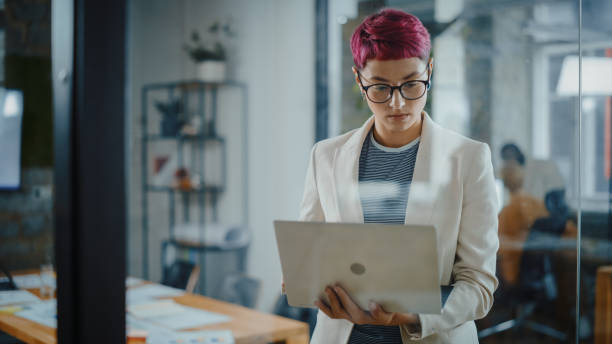 ufficio moderno: ritratto di bella specialista autentica con i capelli rosa corti in piedi, tenendo in mano il computer portatile, concentrato sul suo lavoro. lavorare su design, analisi dei dati, strategia del piano - nonconforming foto e immagini stock