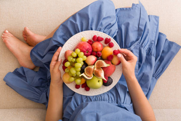 top view on woman sitting cozy on the couch and enjoying delicious summer fruit and berries. girl holding white plate with apricots, raspberries, peaches and figs on her knees. healthy eating. - nature human pregnancy color image photography imagens e fotografias de stock