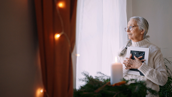 Mujer mayor nostálgica sosteniendo el marco de la foto, de pie junto a la ventana en casa. photo