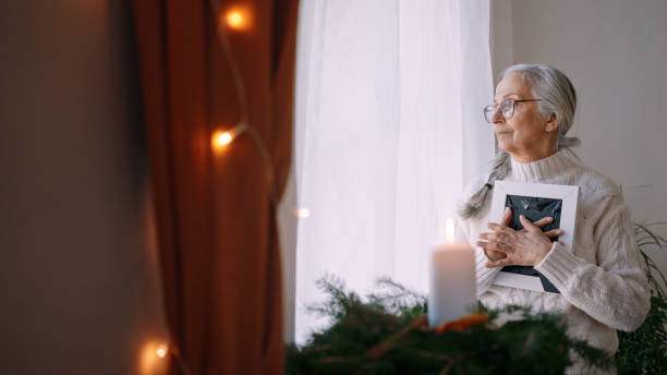 femme âgée nostalgique tenant un cadre photo, debout près de la fenêtre à la maison. - veuve photos et images de collection