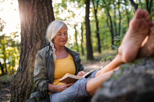 A senior woman relaxing and reading book outdoors in forest.