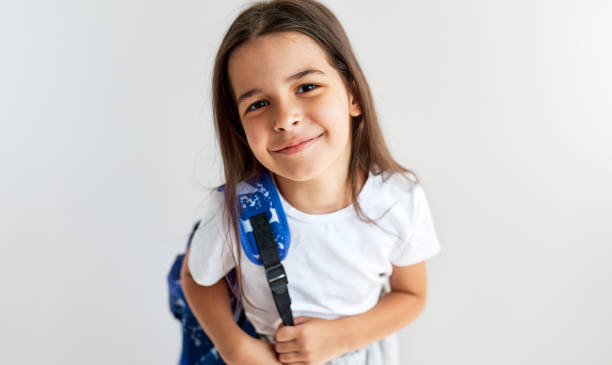 Cute schoolgirl smiling wearing white t-shirt with blue backpack looking at the camera posing against white background. Positive schoolkid with backpack has joyful expression to going back to school. stock photo