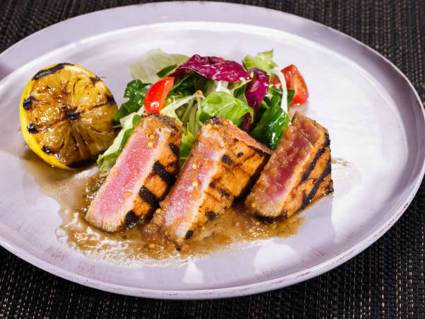 filetto di tonno alla griglia servito con mix di insalata e limone grigliato su un piatto su sfondo scuro - tuna steak fillet food plate foto e immagini stock