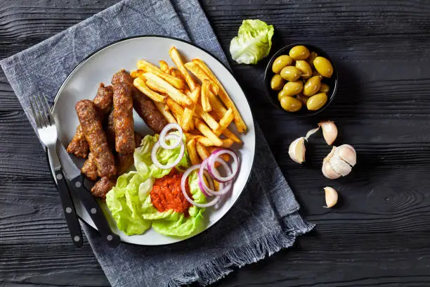 cevapcici, grilled balkan sausages with potato fries, red onion rings, ajvar and fresh lettuce leaves on a plate on a black wooden table with green olives, flat lay, close-up, free space
